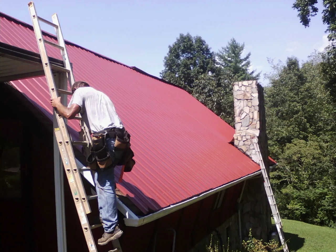Roofer on a ladder beside a red metal roof and stone chimney — Soffit & Fascia Repair by Roofing Brothers in Dover